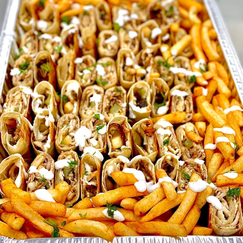 A catering tray filled with rows of SK Shawarma wraps and seasoned french fries, drizzled with white sauce.