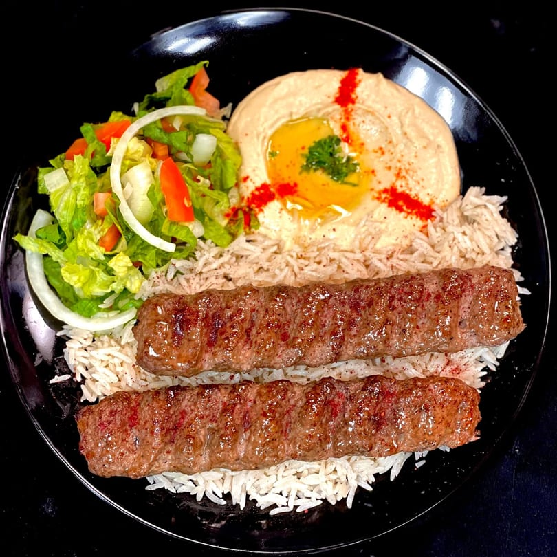 Two grilled kebabs on a bed of rice with a side of hummus and a fresh salad.