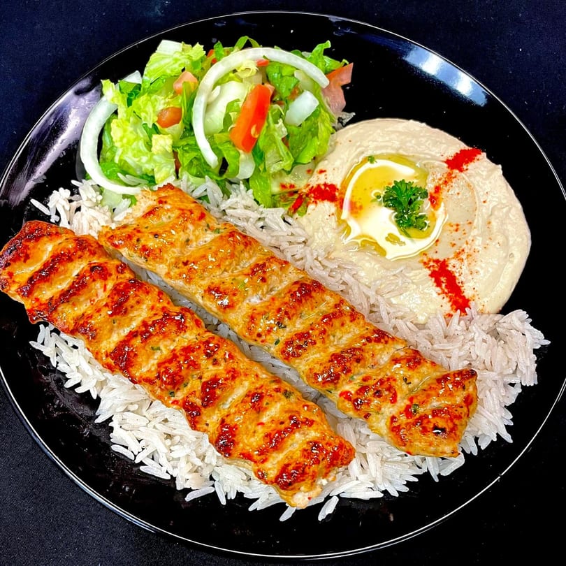 Two grilled chicken skewers on rice with a side of hummus, salad, and onions.